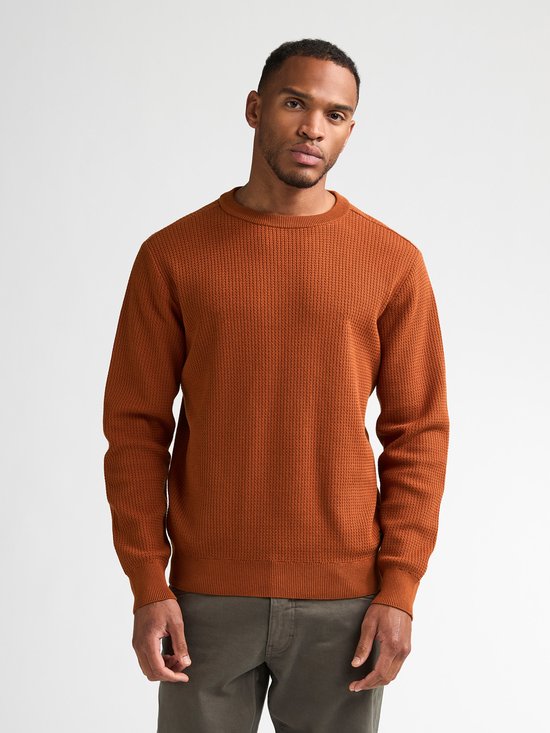 Man van achteren gezien, draagt een oranje trui met ronde hals en wafelpatroon en een grijze broek.