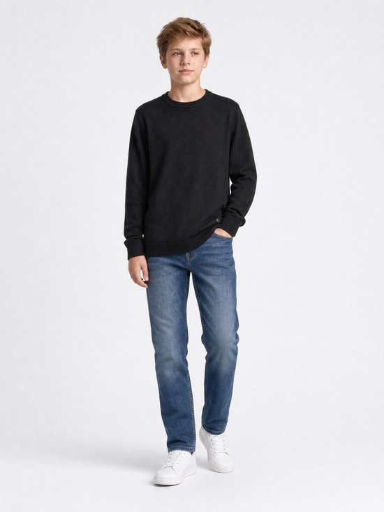 Jongen draagt een zwarte fijngebreide trui, blauwe jeans en witte sneakers.