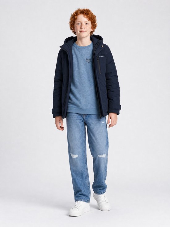 Jongen met krullend rood haar, draagt een donkerblauwe jas, blauwe trui en spijkerbroek.
