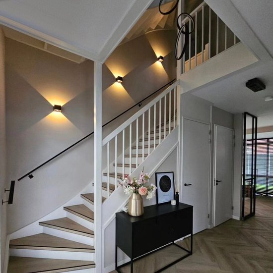 Cage d'escalier avec marches en bois, balustrade blanche et quatre appliques murales noires projetant une lumière chaude sur le mur.