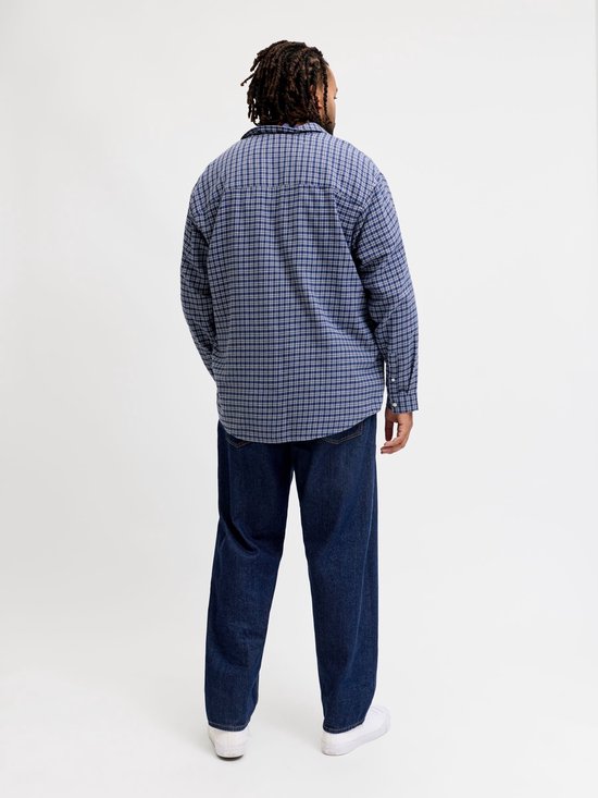 Man met dreadlocks draagt een blauw geruite blouse en donkere jeans.