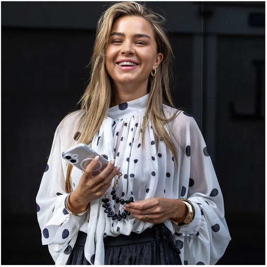 Vrolijke vrouw met witte blouse met polkadot print, zwarte rok, houdt transparante telefoonhoes met hartjes polsband vast.