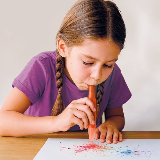 Een meisje met vlechten blaast op een oranje pen, waardoor gekleurd poeder op een wit vel papier verschijnt.