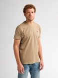 Man met krullend rood haar, staat frontaal, draagt lichtbruin t-shirt met borstlogo en spijkerbroek.
