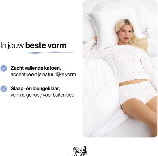 Model liggend op een bed, draagt een witte pyjamaset met rode hartjes.
