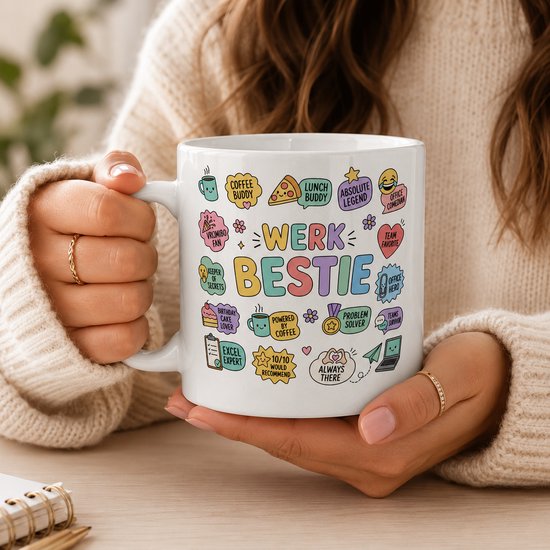 Witte mok met de tekst "WERK BESTIE" en diverse illustraties, vastgehouden door een persoon met beige trui.