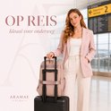 Vrouw met roze blazer en witte broek bij een luchthaven met een roze rugzak op een zwarte koffer.
