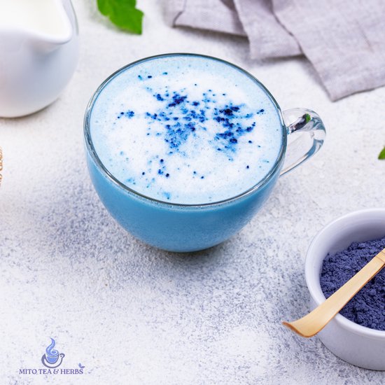 Glazen mok gevuld met een blauwe matcha drank, afgetopt met wit schuim en blauwe poederdeeltjes, naast een kommetje met blauwe matcha.