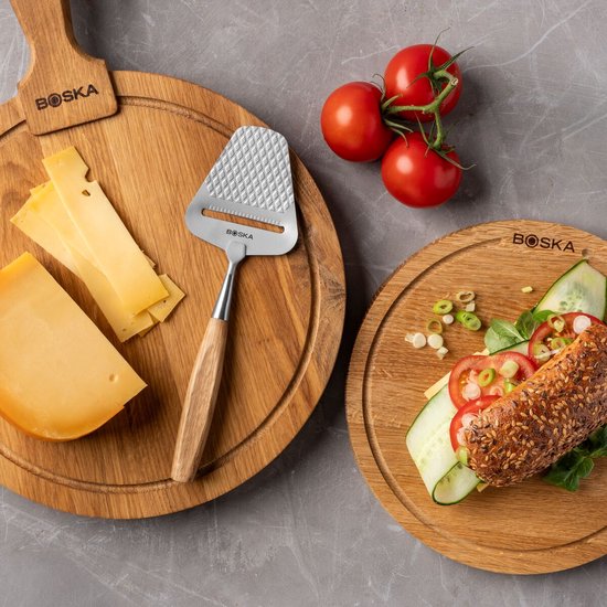 Houten kaasplank met kaasrasp, plakjes kaas en een stuk kaas. Tros tomaten en een broodje op een aparte plank.