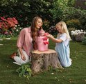 Vrouw en meisje zitten in tuin bij boomstronk. Meisje geeft Luvibubs pop in pot water. Pop heeft roze haar en blaadjes.