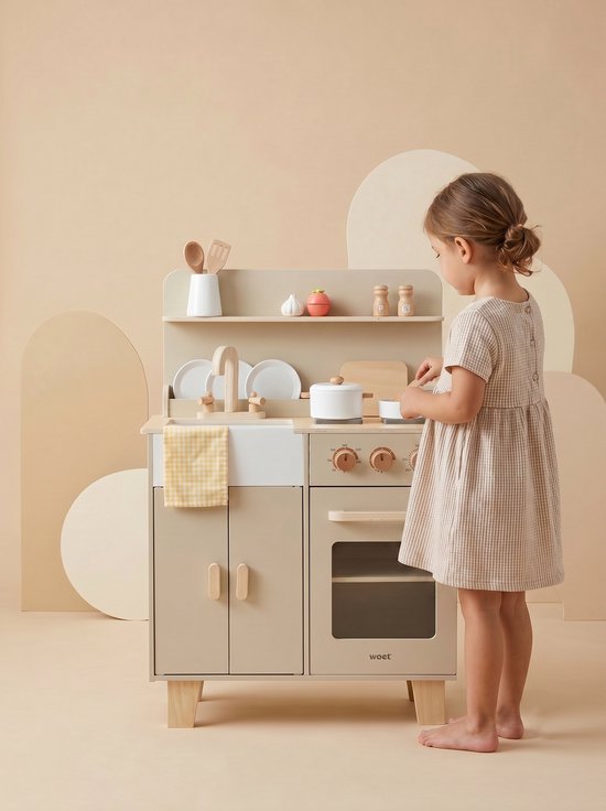 Meisje in beige geruite jurk speelt met beige houten keuken. De keuken heeft witte spoelbak, fornuis, magnetron en accessoires.