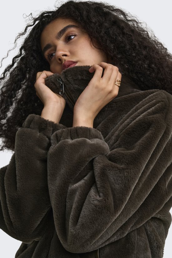 Close-up van een model met krullend haar die de kraag van een donkerbruine teddy jas optrekt.
