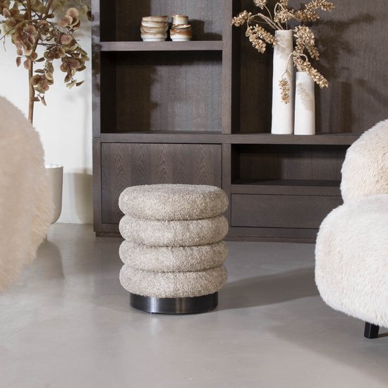 Gestapelde taupe bouclé poef met een donkere metalen voet, tussen witte vazen en een fluffy fauteuil.
