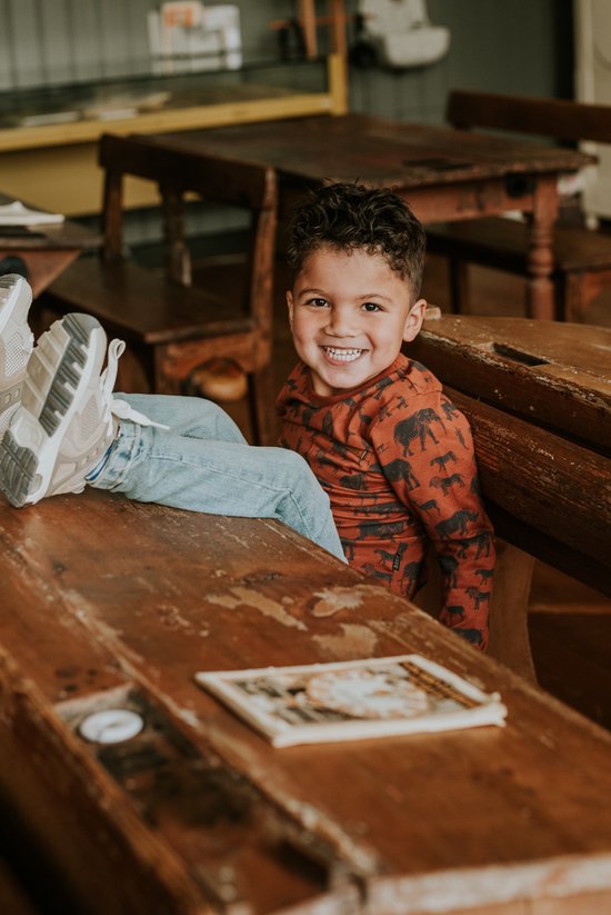 Jongetje in oranje shirt met dierenprint, zittend op een houten schoolbank.