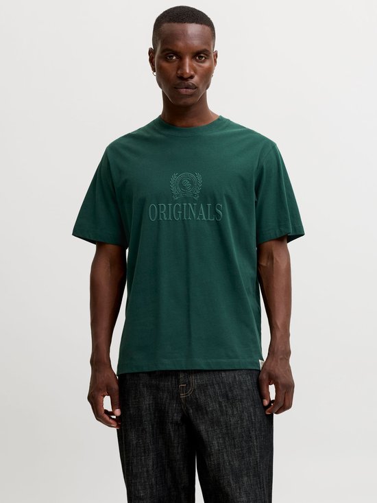 Model draagt een donkergroen t-shirt met ronde hals en reliëf 'ORIGINALS' print op een donkergrijze broek.
