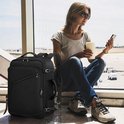 Zwarte reistas naast een vrouw met zonnebril die bij een raam zit op een vliegveld, met telefoon en koffie.