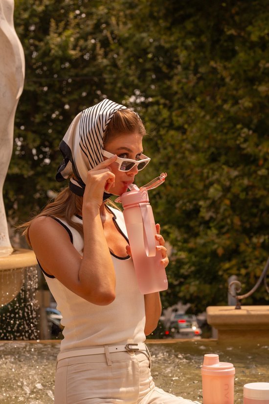 Een vrouw met een sjaal en zonnebril drinkt uit een roze Cabau fles bij een fontein.