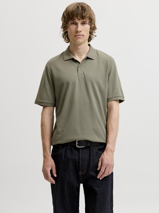 Man met de voorkant naar de camera, draagt een kaki poloshirt met korte mouwen en donkere jeans.