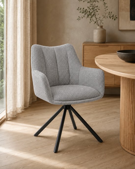 Grijze eetkamerstoel met armleuningen, een stoffen bekleding en een zwart metalen draaipoot, geplaatst naast een houten tafel.