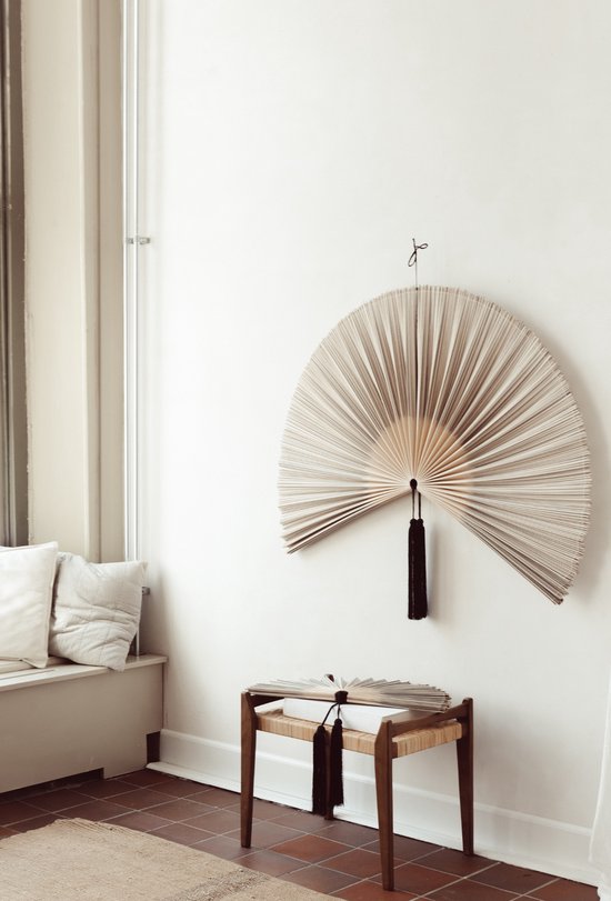 Een decoratieve waaier met zwarte kwast hangt aan de muur boven een houten bankje met een waaier.