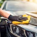 Een hand met een zwarte handschoen poetst met een gele microvezeldoek over de glimmende zwarte motorkap van een auto.