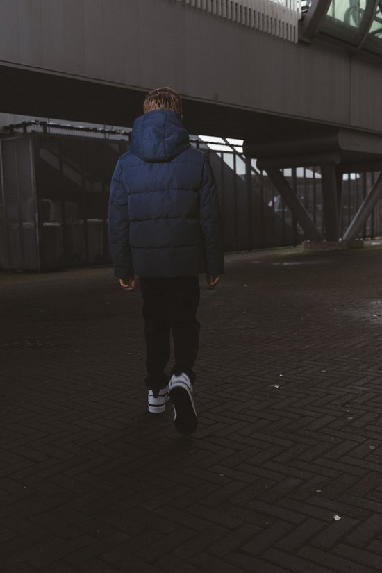 Jongen in een donkerblauwe pufferjas met capuchon en zwarte broek, lopend weg van de camera.