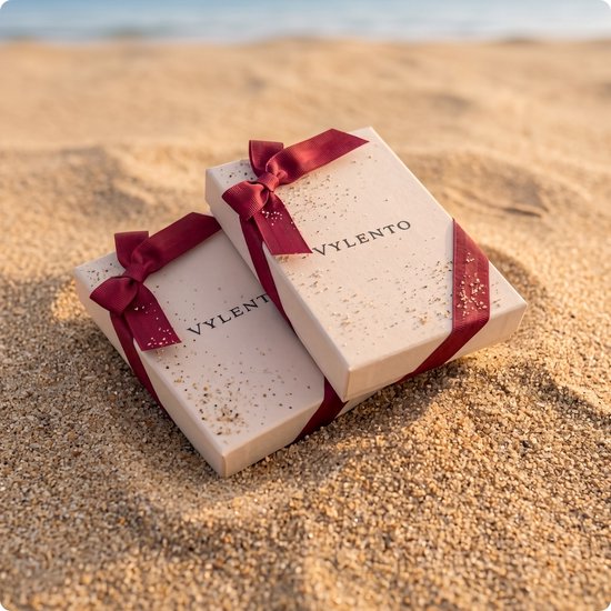 Deux boîtes cadeaux beiges avec des rubans bordeaux et des éclats dorés, sur une plage ensoleillée.
