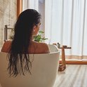 Vrouw met lang, donker, nat haar ontspant in een wit bad met een lichte achtergrond.