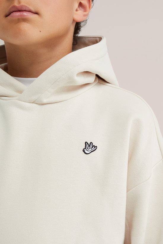 Close-up van een jongen in een beige hoodie met een klein, geborduurd handlogo.