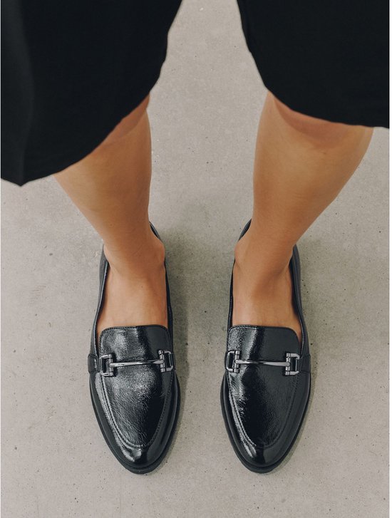 Een vrouw draagt zwarte lakleren loafers met zilverkleurige gespen.