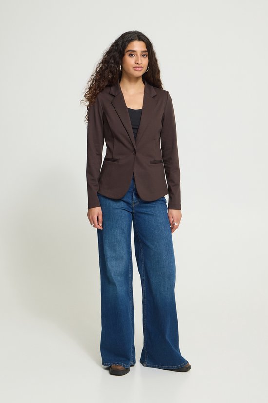 Vrouw draagt een bruine blazer met een zwarte top en wijde spijkerbroek. Ze heeft krullend haar en grote oorbellen.