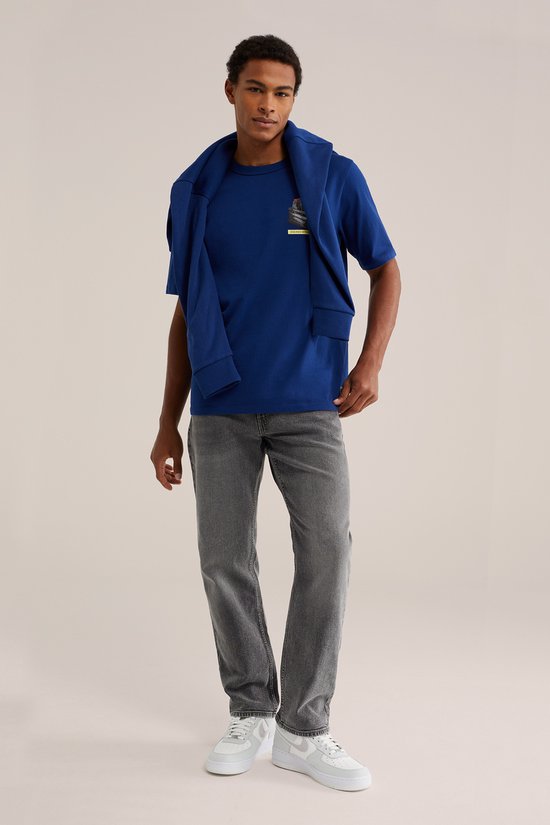 Model staat frontaal, draagt blauwe T-shirt met print en grijze jeans. Blauwe trui over de schouders.