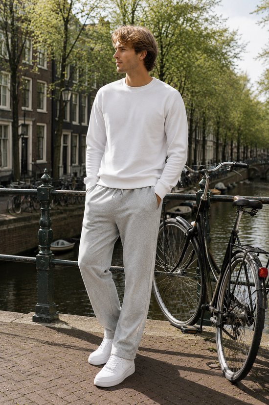 Een man met blond haar staat buiten met de handen in zijn zakken van een grijze joggingbroek en draagt een witte sweater.