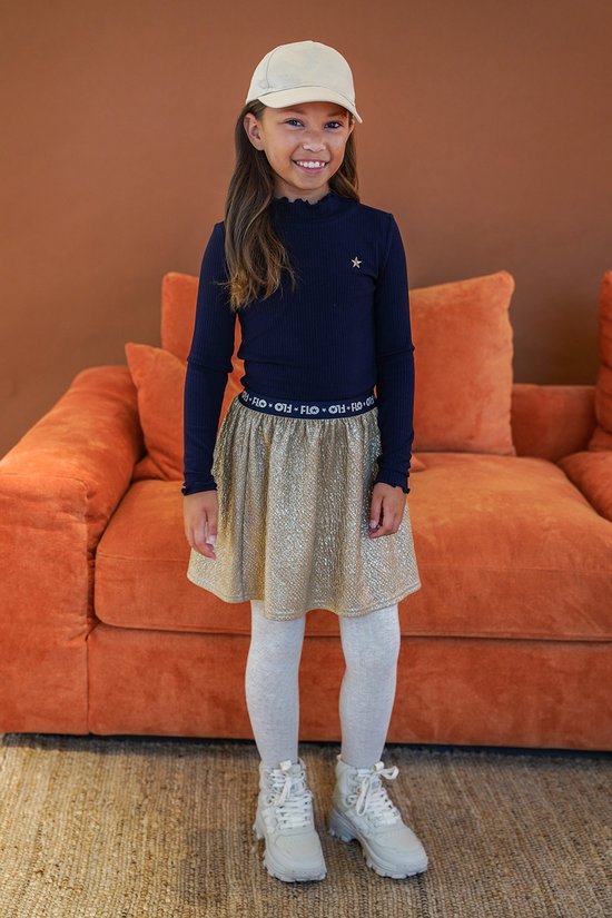 Meisje met bruin haar en een pet draagt een donkerblauw ribgebreid shirt met ster en een glimmende gouden rok met "Like Flo" tekst.