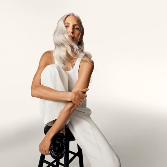 Model met lang, grijs haar draagt een witte tanktop en witte broek, zittend op een kruk, met zwarte loafers.