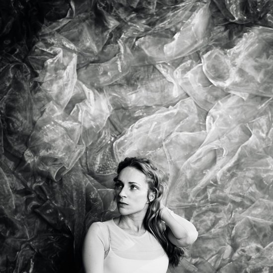 Zwart-wit portret van Agnes Obel met golvend haar, haar hand in haar nek, tegen een achtergrond van verfrommeld materiaal.