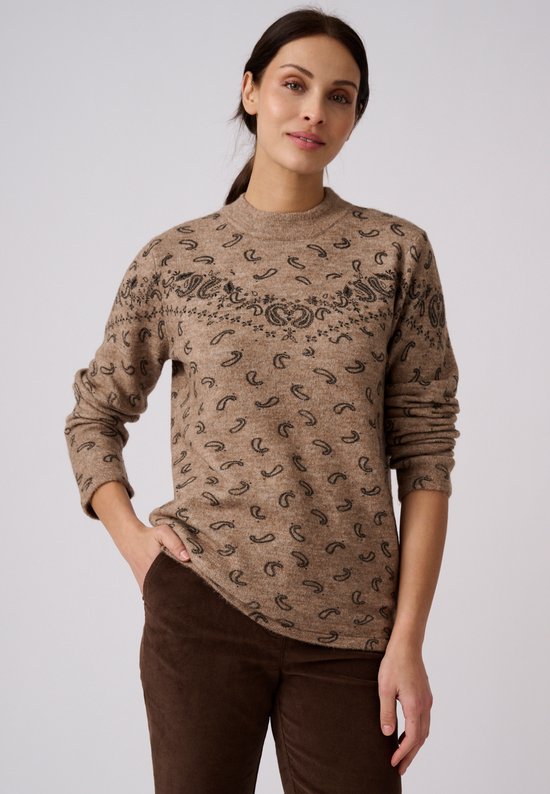 Model draagt een beige trui met zwarte paisley-details en een ronde hals, één hand in de zak van een bruine broek.