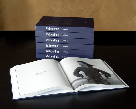 Boek 'Welkom thuis' met opengeslagen pagina met een zwart-witfoto van een vrouw. Titel en naam 'Henk Warrink' op de ruggen van gestapelde boeken.
