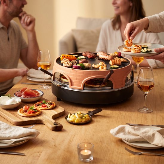 Een gezelschap eet aan een houten tafel rondom een raclette en grill met diverse gerechten.