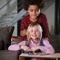 Jongen met afro-kapsel en meisje met blond haar spelen IQ Fit spel aan een tafel. Het meisje heeft de stukken in haar handen.