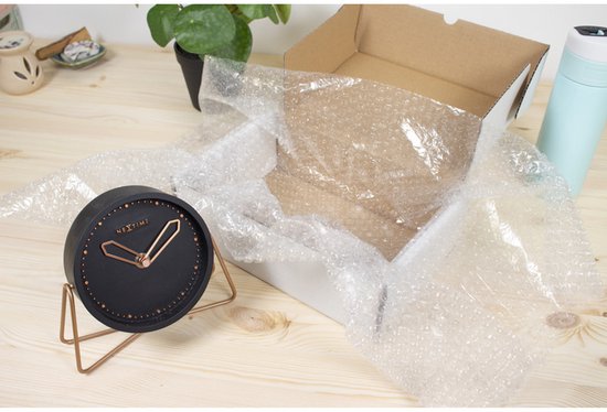 Een zwart, rond horloge met koperkleurige wijzers op een standaard, naast een geopende kartonnen doos met noppenfolie.