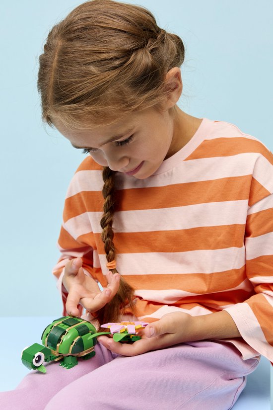 Een meisje met een vlecht en een oranje gestreepte trui houdt een groene legoschildpad met een roze bloem vast.