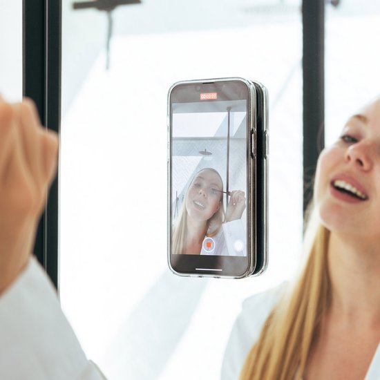 Vrouw filmt zichzelf in de spiegel terwijl ze make-up aanbrengt, haar telefoon hangt aan de spiegel.