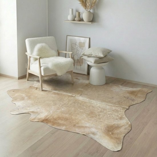 Beige koeienhuid vloerkleed in een modern ingerichte woonkamer, liggend op een houten vloer voor een witte fauteuil.