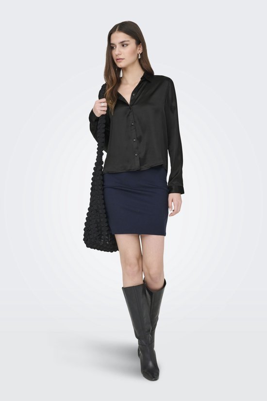 Model draagt zwarte satijnen blouse met knoopjes, donkerblauwe rok en zwarte laarzen.