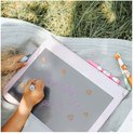 Kind tekent 'JEANNE' met hartjes op grijs lichttablet, buiten op kleed.