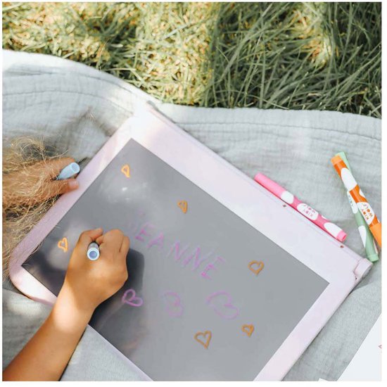 Kind tekent 'JEANNE' met hartjes op grijs lichttablet, buiten op kleed.