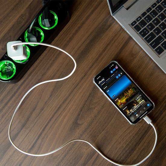 Zwarte stroomstrip met vier groene, verlichte stopcontacten, een smartphone die oplaadt via USB en een laptop op een houten tafel.