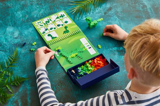 Kind bouwt een groene dinosaurus met Plus Plus bouwstenen. Instructies op openklapbare drukker.