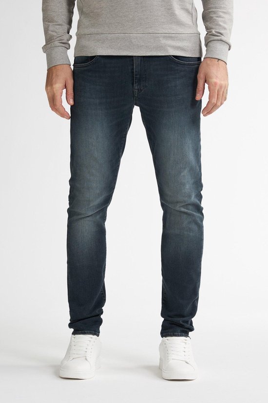 Man met donkerblauwe slim fit jeans, licht vervaagd aan de voorkant, met witte sneakers.